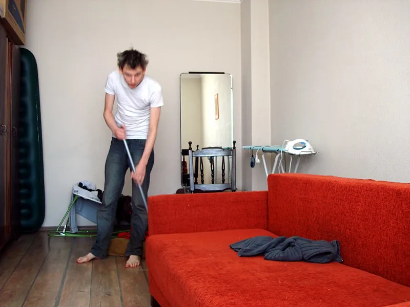 Un jeune homme passe l’aspirateur au pied d’un canapé en tissu rouge