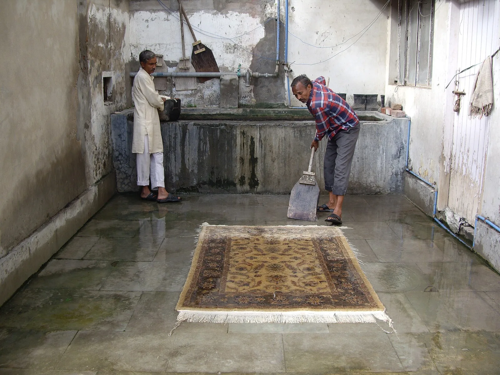 Deux hommes lavent un tapis à grande eau dans un atelier de nettoyage