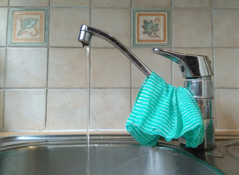 L’eau coule du robinet dans un évier inox, un torchon sur le mitigeur