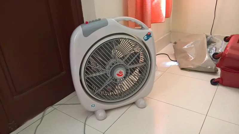 Un ventilateur posé au sol, tourné vers la pièce, près d’une fenêtre